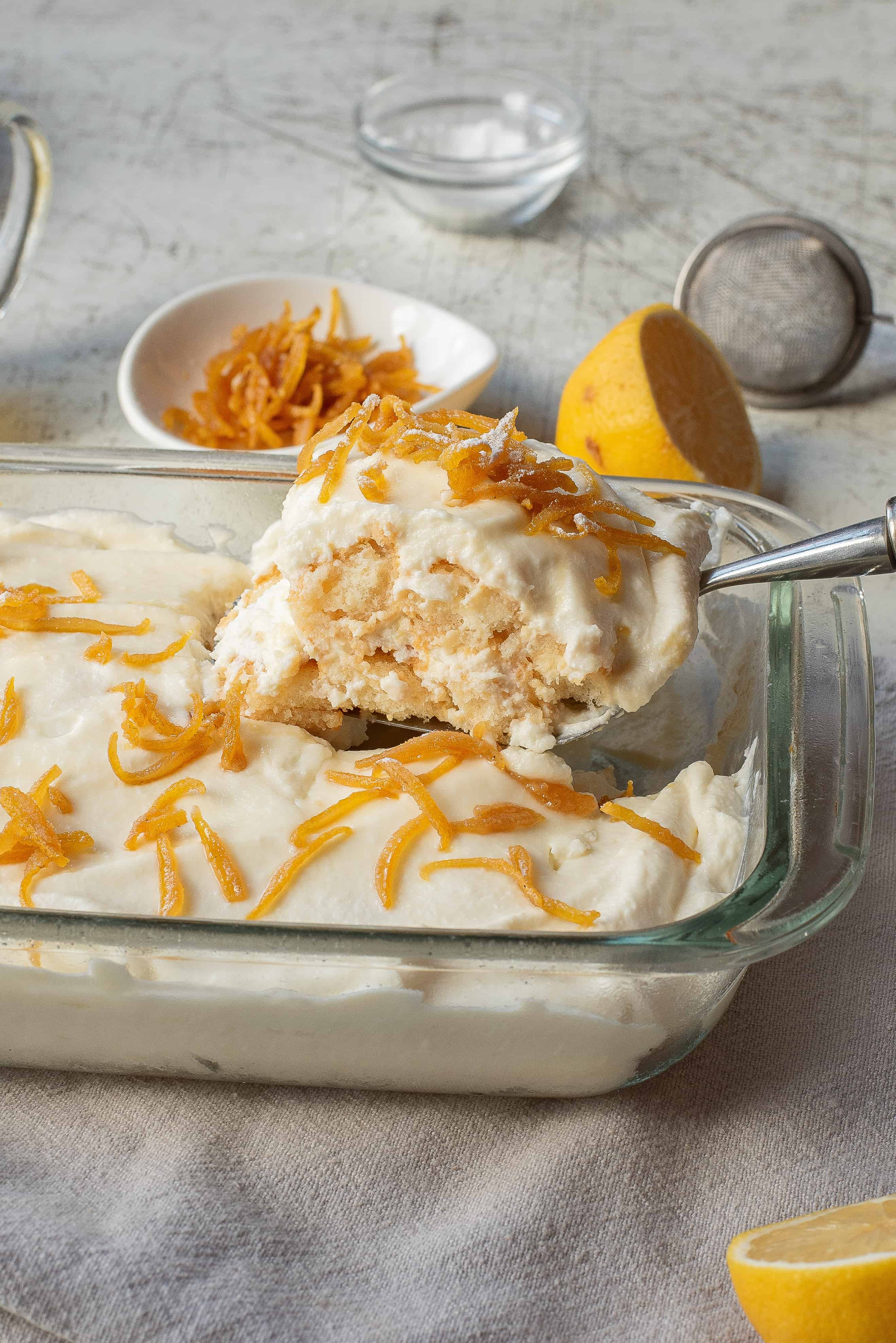 A glass baking dish filled with a creamy dessert topped with orange zest. A serving spoon has lifted a portion, revealing a layered texture. A small plate of orange zest and a halved lemon are in the background.