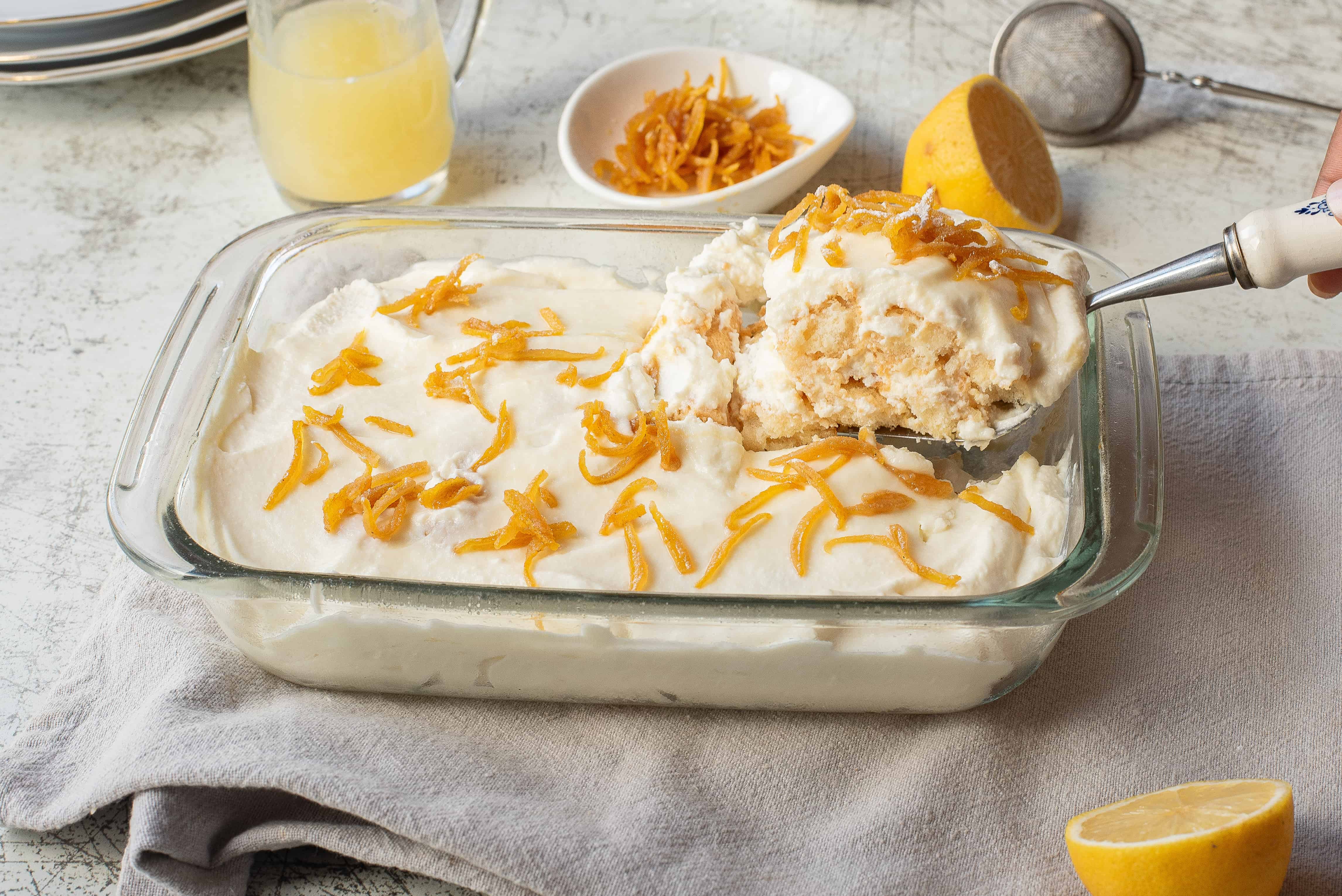 A glass baking dish contains a creamy dessert topped with lemon zest. A large serving spoon holds a portion above the dish. A half lemon, a bowl of zest, and a pitcher are nearby on a light surface.
