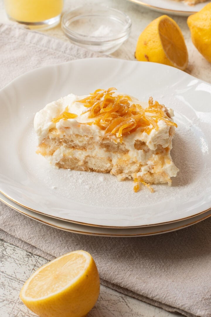A slice of lemon dessert with creamy layers and candied lemon peel on top is served on a white plate, with fresh lemons, a napkin, and a glass bowl in the background.