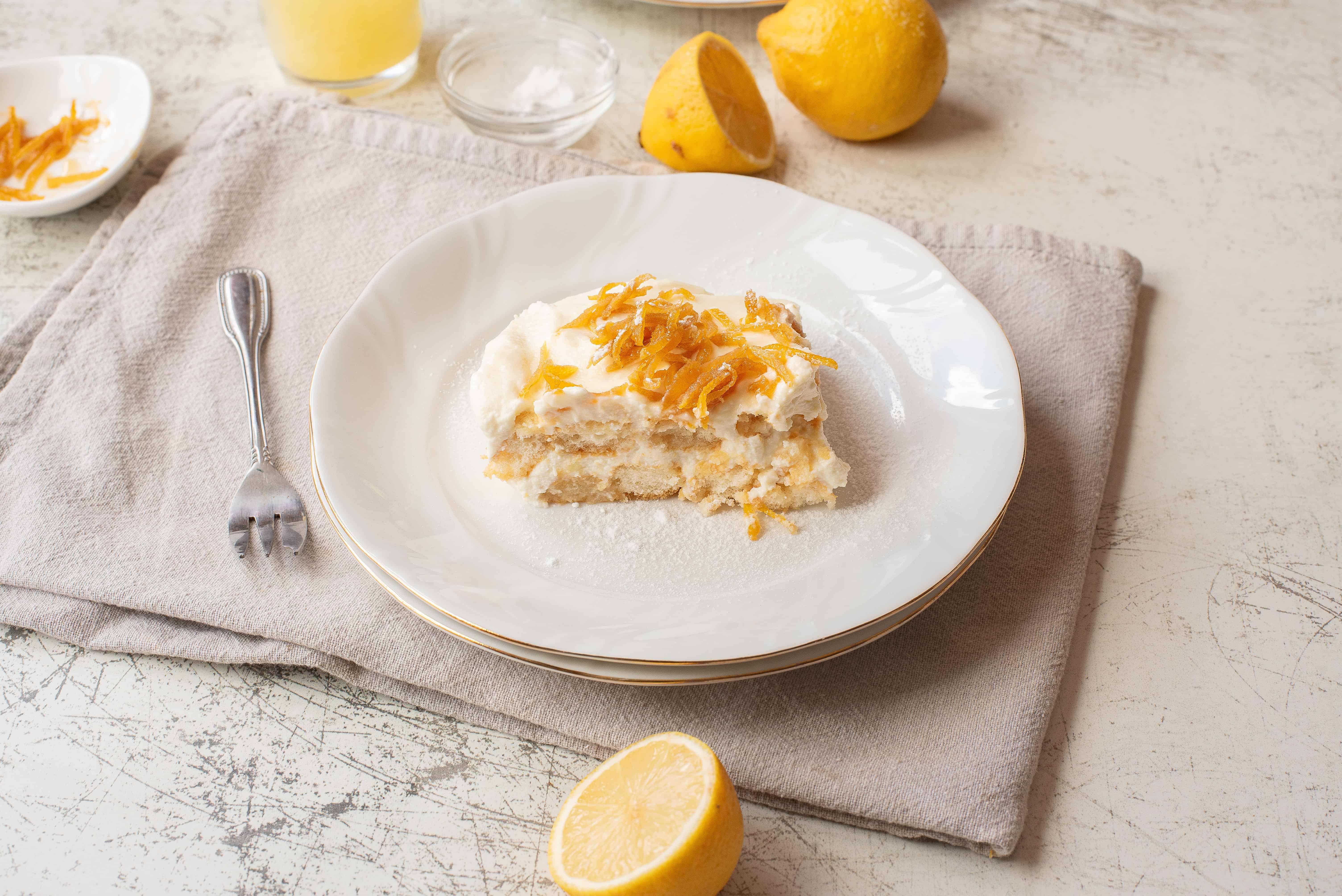 A slice of lemon dessert topped with candied lemon peel sits on a white plate with a fork beside it. Whole and halved lemons and small bowls with ingredients are in the background on a beige cloth.