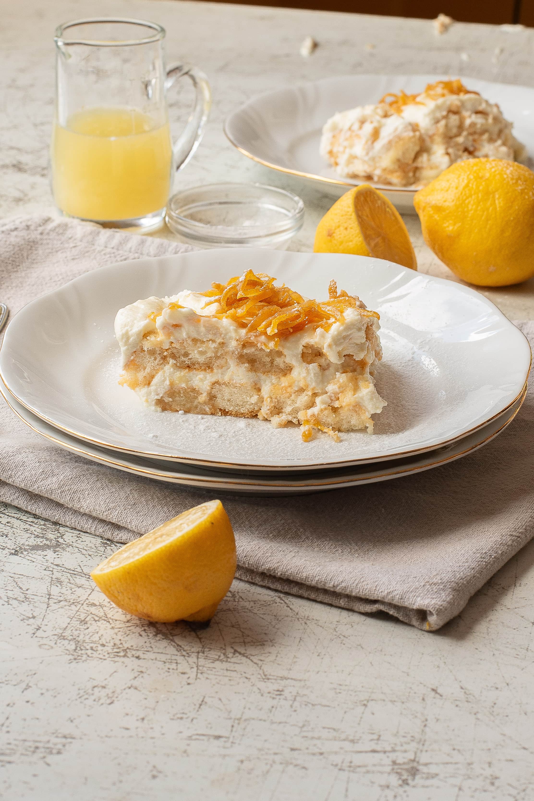 A slice of lemon dessert topped with whipped cream and zest on a white plate, next to a halved lemon. In the background, another plate with dessert, whole lemon, and a glass pitcher with yellow liquid are visible.