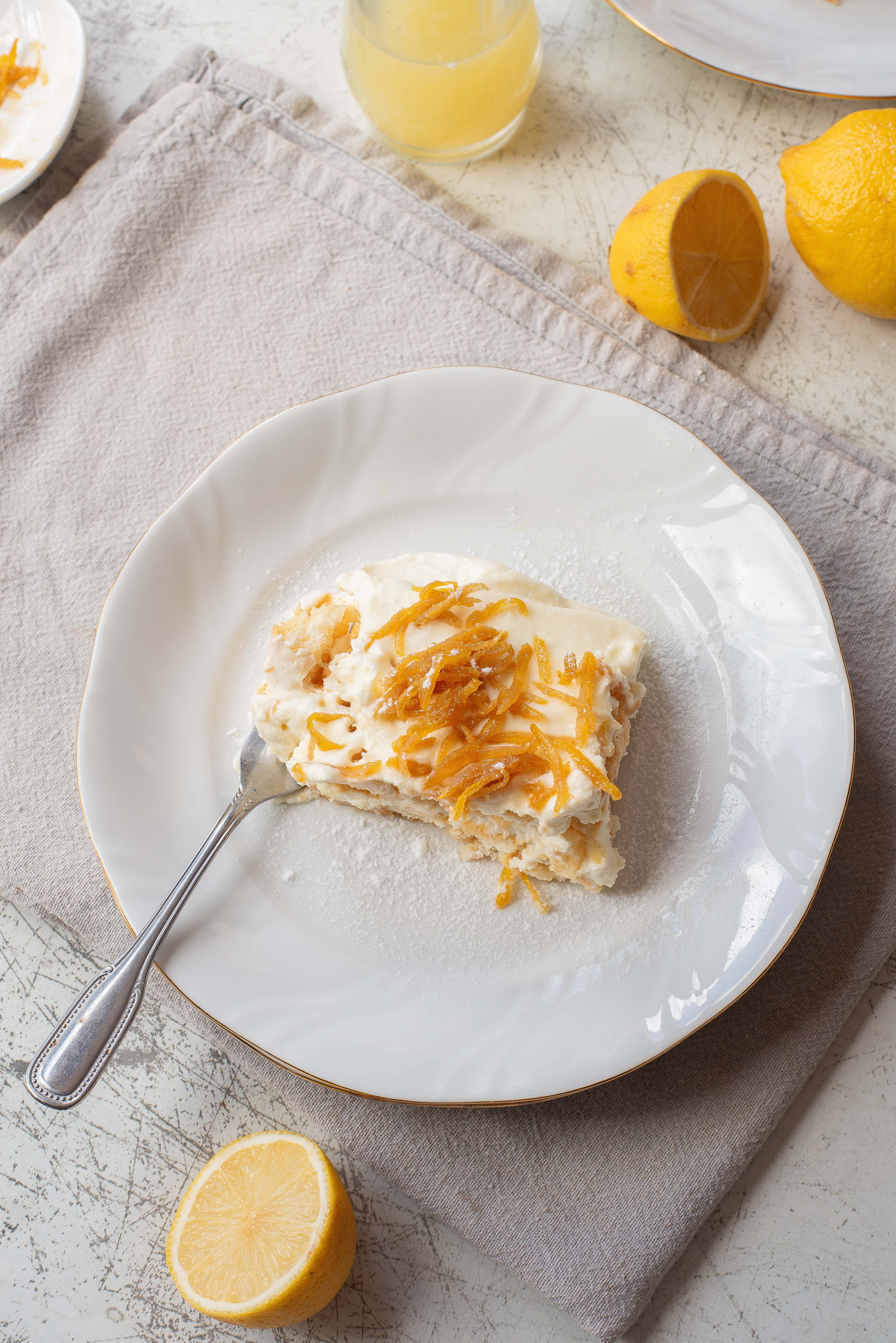 A white plate with a slice of lemon cake topped with cream and lemon zest sits on a beige cloth. A fork rests on the plate. Nearby are halved lemons and a glass of juice on a light-textured surface.