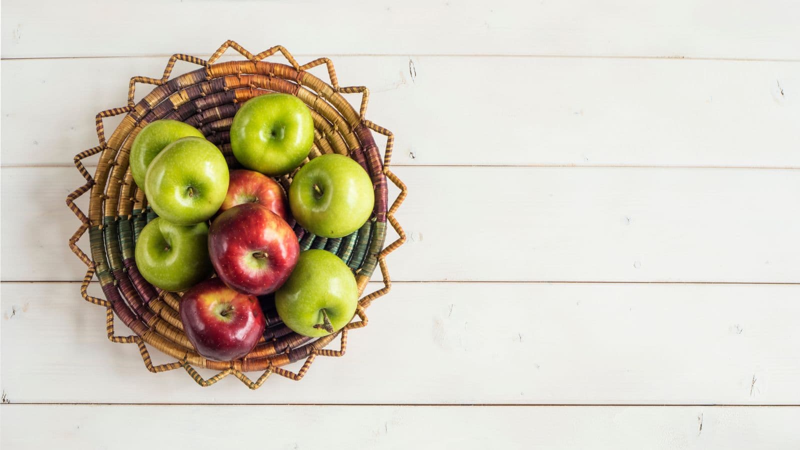 A woven plate filled with five green apples and three red apples sits on a white wooden surface. The plate has a zigzag-patterned border in earthy tones. The right side of the image is empty.