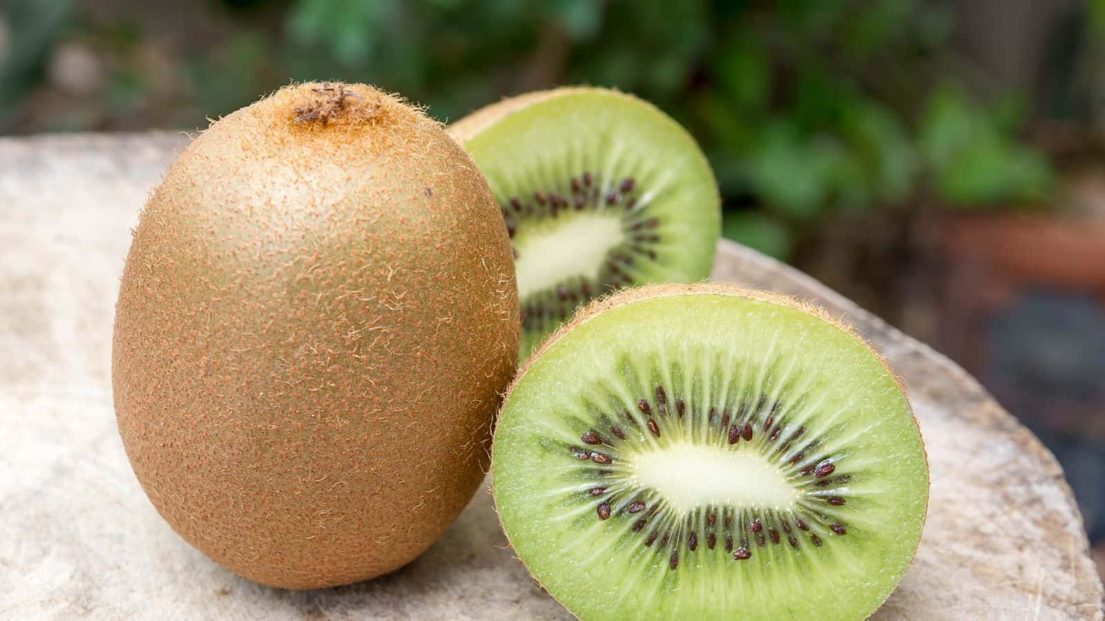 A whole kiwi fruit and a kiwi fruit cut in half are placed on a light-colored surface. The inside of the kiwi is bright green with small black seeds, while the outer skin is brown and fuzzy.