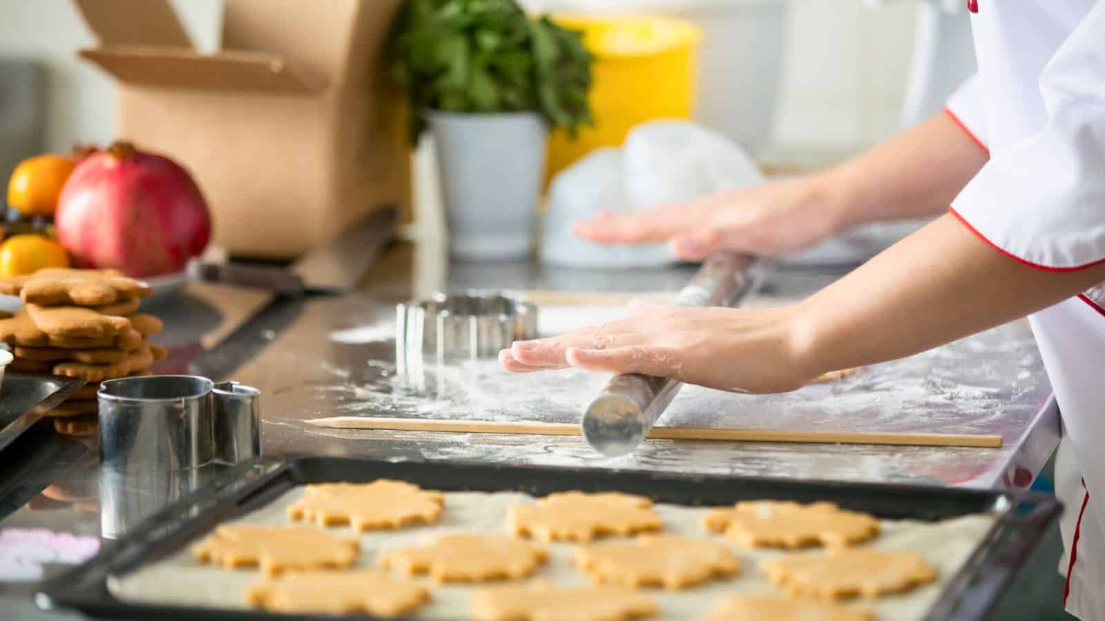 A person in a white coat rolls out dough with a rolling pin on a floured surface. Cookie cutters and shaped cookies are on the table, with a baking tray in the foreground and ingredients in the background.