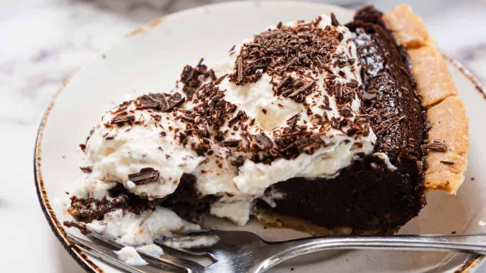 A slice of chocolate pie with a flaky crust topped with whipped cream and chocolate shavings sits on a plate with a fork.