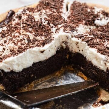 A chocolate cream pie in a metal pie tin, topped with whipped cream and chocolate shavings. A slice has been cut and a metal pie server is visible under the empty space.