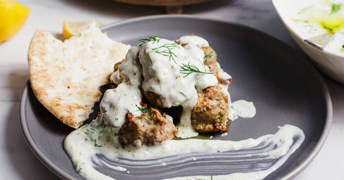 A gray plate holds several Greek Turkey Meatballs with Feta, topped with creamy white sauce and fresh dill, next to a folded piece of pita bread. A smear of sauce is also on the plate, with a bowl and lemon wedge in the background.