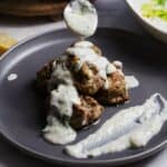A gray plate holds several Greek Turkey Meatballs with Feta stacked together, covered with a creamy white sauce. A spoon is drizzling more sauce on top, and a smear of sauce is spread on the side of the plate.