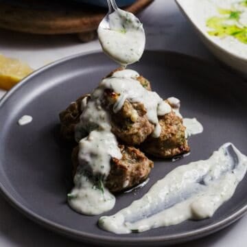 A gray plate holds several Greek Turkey Meatballs with Feta stacked together, covered with a creamy white sauce. A spoon is drizzling more sauce on top, and a smear of sauce is spread on the side of the plate.
