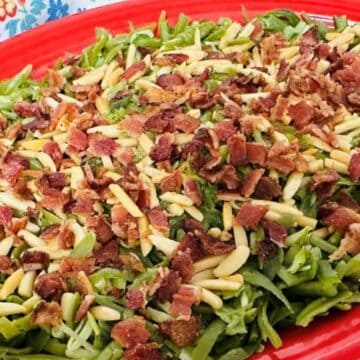 A red oval serving dish filled with green beans topped with crispy bacon pieces and slivered almonds, with a serving fork beside the dish on a wooden surface.