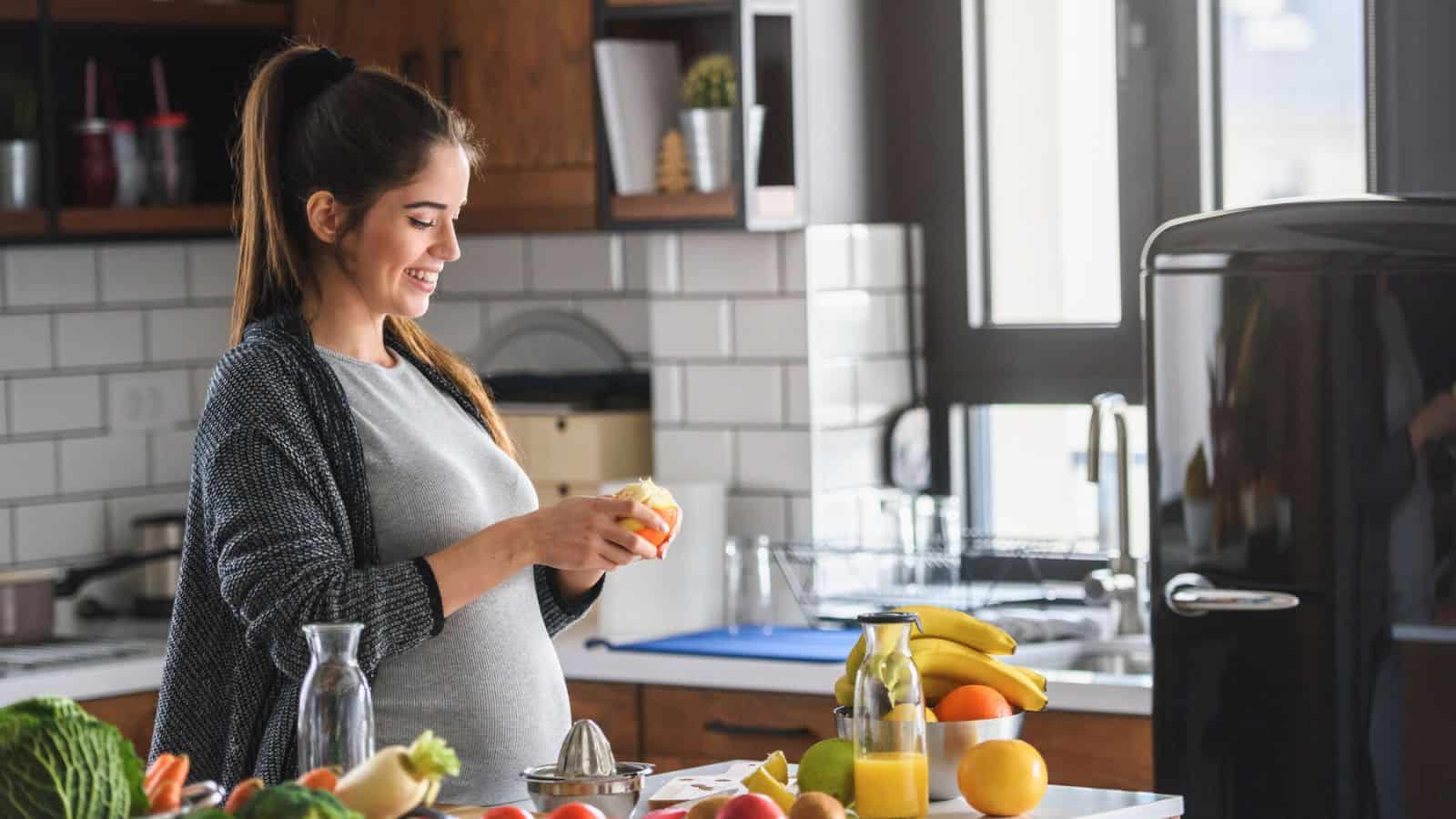 A pregnant woman stands in a kitchen, smiling while peeling an orange. There are various fruits, vegetables, and kitchen items on the counter around her. Natural light comes in through a window.