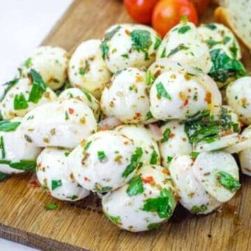 A pile of marinated mozzarella balls garnished with chopped herbs and spices sits on a wooden board, with cherry tomatoes and a slice of bread in the background.