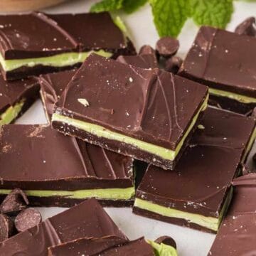 Rectangular pieces of chocolate mint bars are stacked on a surface surrounded by chocolate chips and mint leaves. Each bar has a dark chocolate top and bottom layer with a green mint layer in the middle.