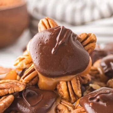 A close-up of chocolate pecan turtles, each made with pecans, caramel, and topped with smooth chocolate, piled together on a surface with a bowl and striped cloth in the background.