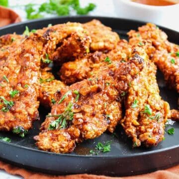 A black plate filled with several crispy, golden-brown chicken tenders garnished with chopped green herbs, placed on a brown cloth with a bowl of dipping sauce in the background.