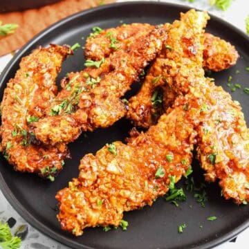 A black plate with several crispy, golden-brown chicken tenders coated in sauce and garnished with chopped herbs, placed on a white speckled surface with parsley scattered around.