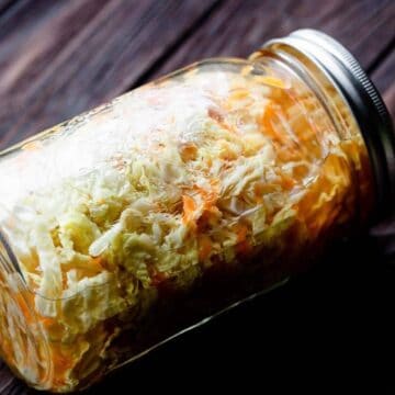 A glass mason jar filled with shredded cabbage and carrots, placed on its side on a dark wooden surface. The jar is sealed with a metal lid.