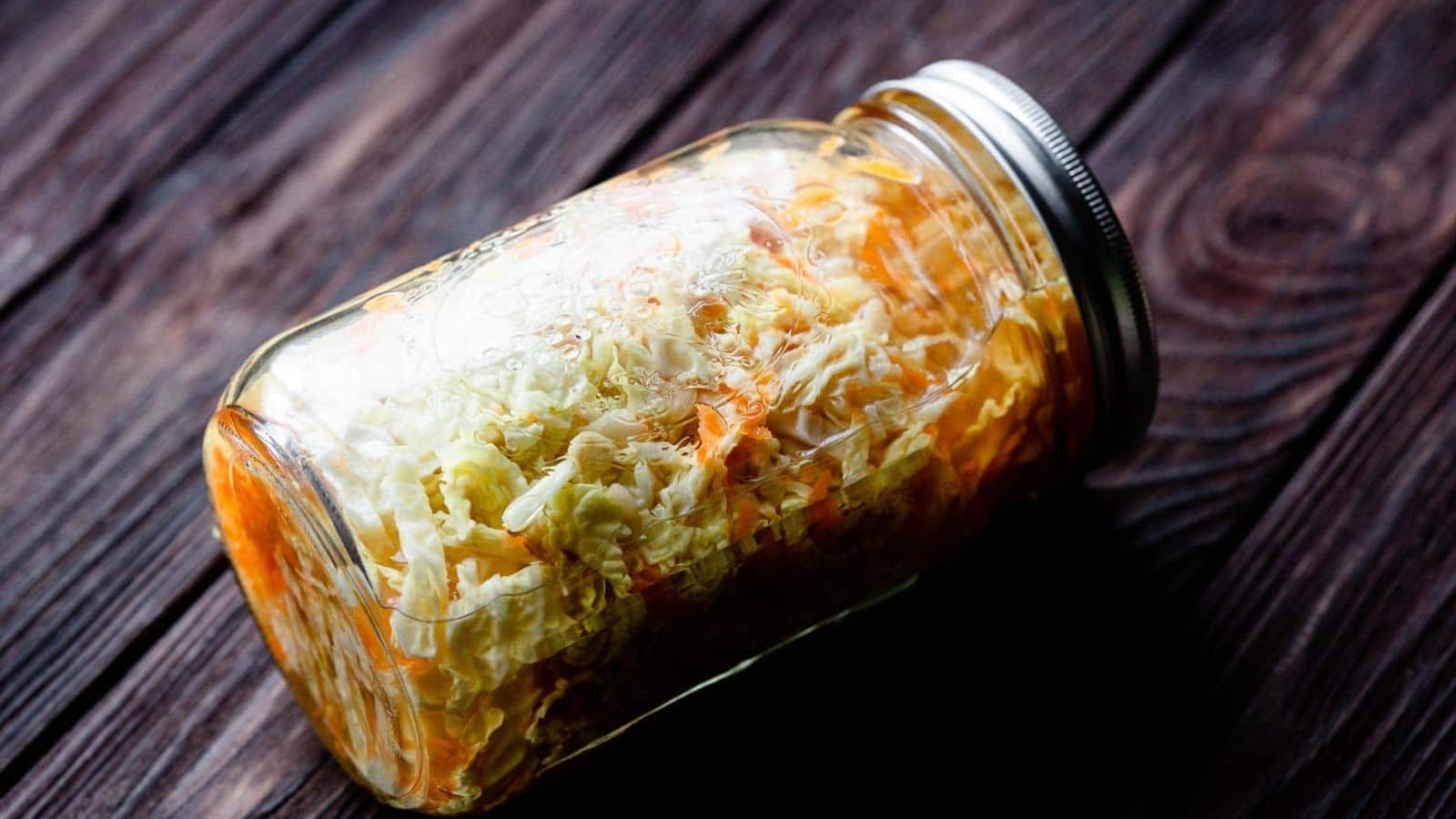 A glass mason jar filled with shredded cabbage and carrots, placed on its side on a dark wooden surface. The jar is sealed with a metal lid.