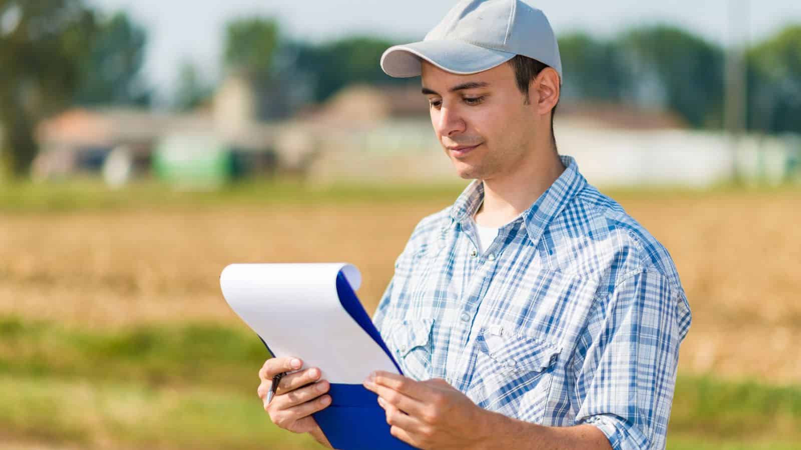 A person wearing a light gray cap and a plaid shirt stands outdoors in a field, holding and reading from a clipboard with papers. The background shows blurred fields and trees.