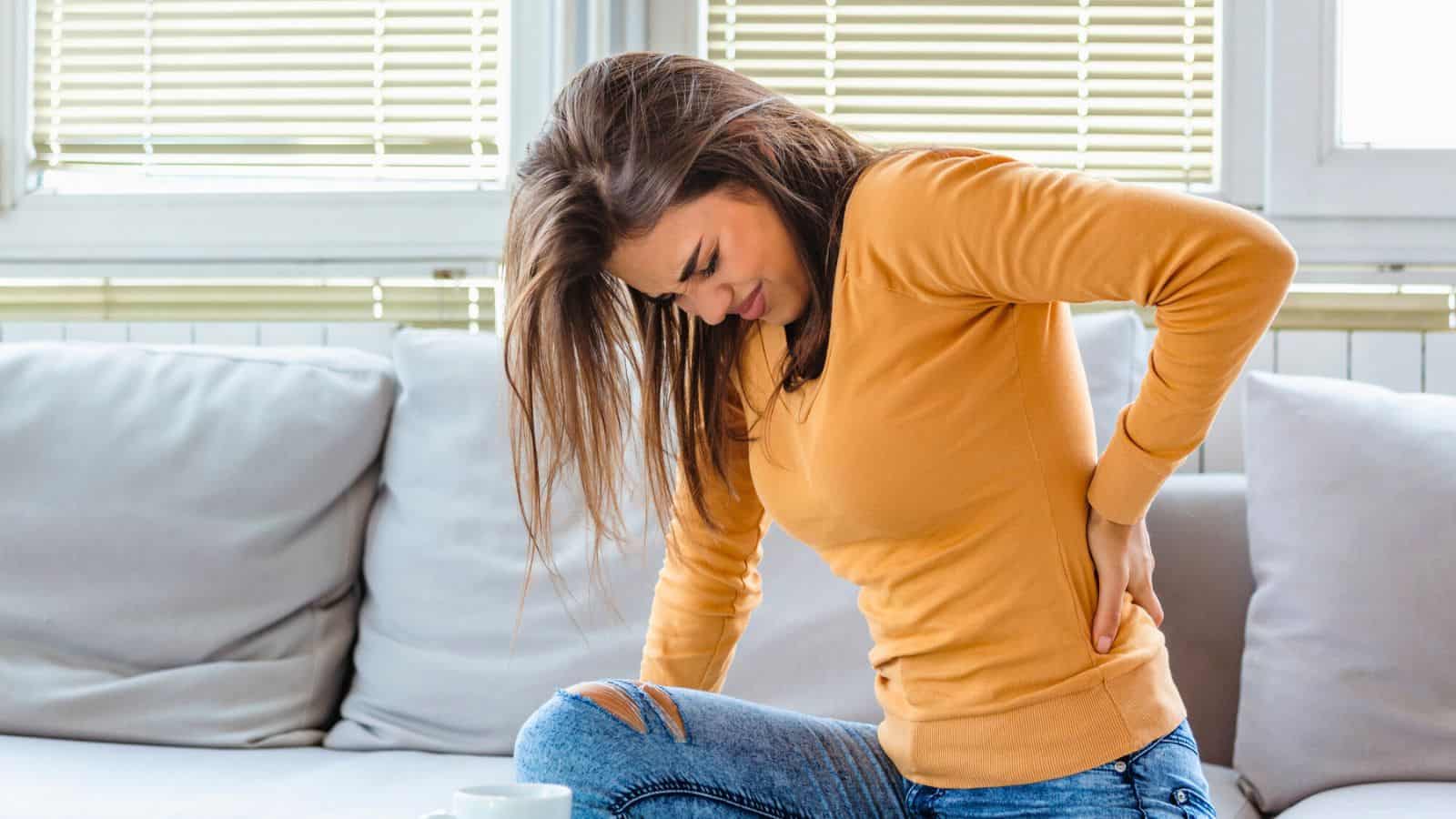A woman sitting on a couch holds her lower back with one hand and appears to be in pain. She is wearing a mustard yellow sweater and ripped blue jeans. Blinds cover the windows in the background.