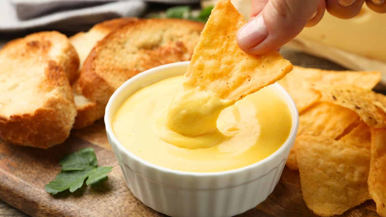A hand dips a triangular tortilla chip into a small white bowl of melted cheese sauce. Toasted bread slices, more chips, and a sprig of parsley are on a wooden board nearby.