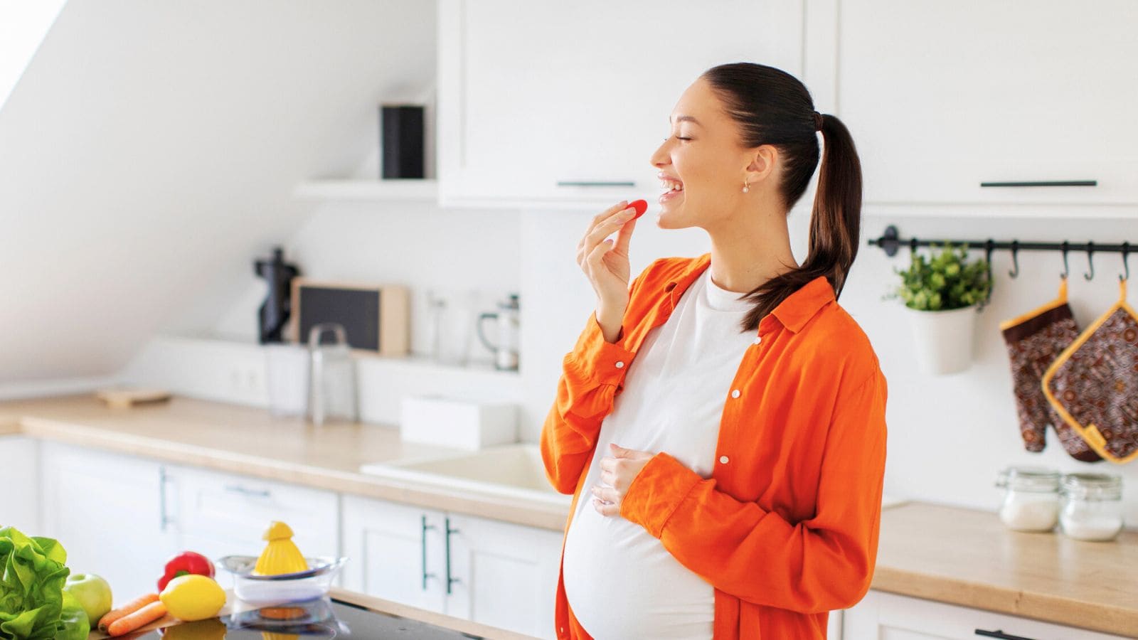 A pregnant woman stands in a bright kitchen, holding her belly with one hand and eating a slice of tomato with the other. Fresh vegetables are on the counter in front of her. She is wearing an orange shirt over a white top.