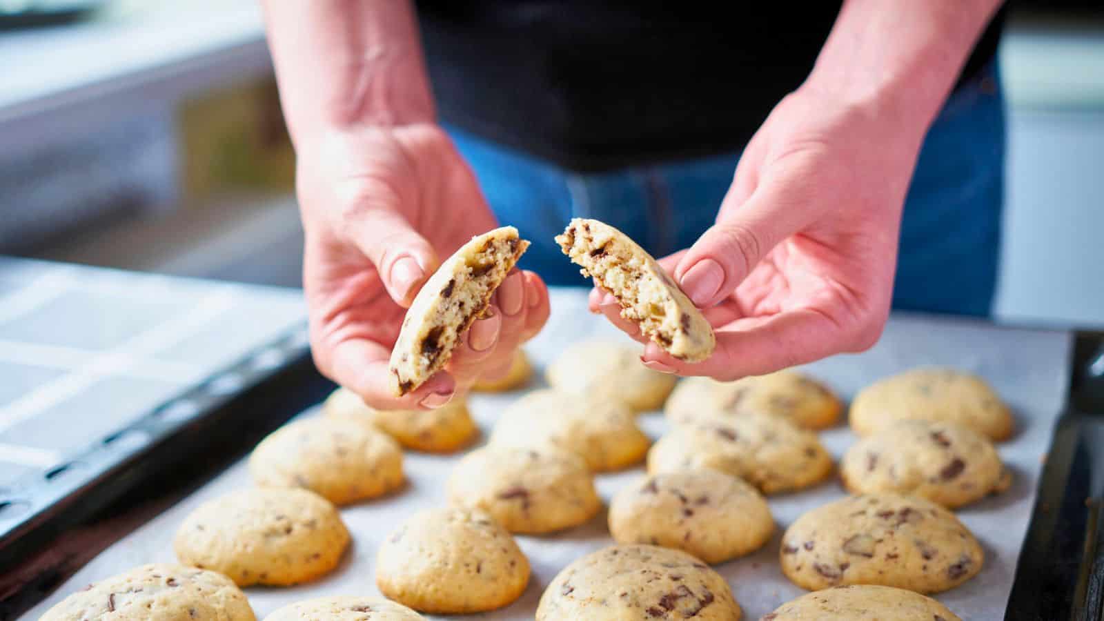 A person holds a chocolate chip cookie broken in half over a baking tray filled with more cookies on parchment paper. The background shows part of a kitchen countertop.