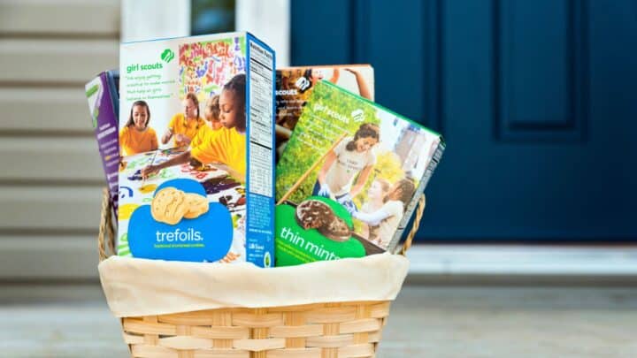 A woven basket containing boxes of Girl Scout cookies, including Trefoils and Thin Mints, is placed on a porch in front of a blue door.