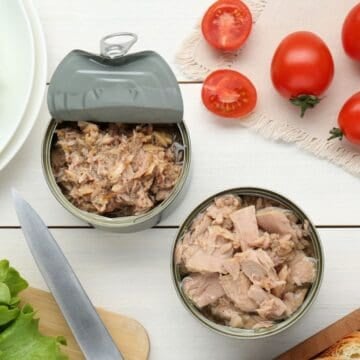 Two opened cans of tuna are on a white table surrounded by fresh lettuce leaves, whole and sliced tomatoes, a knife, a cutting board with bread slices, and a white plate.