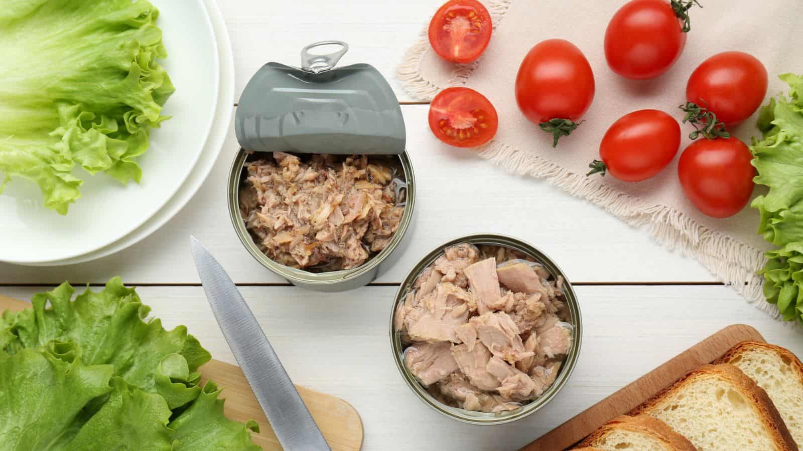 Two opened cans of tuna are on a white table surrounded by fresh lettuce leaves, whole and sliced tomatoes, a knife, a cutting board with bread slices, and a white plate.