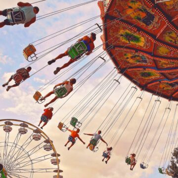 People ride a colorful swing carousel at a fairground, with legs outstretched as the ride spins. A Ferris wheel is visible in the background against a partly cloudy sky.