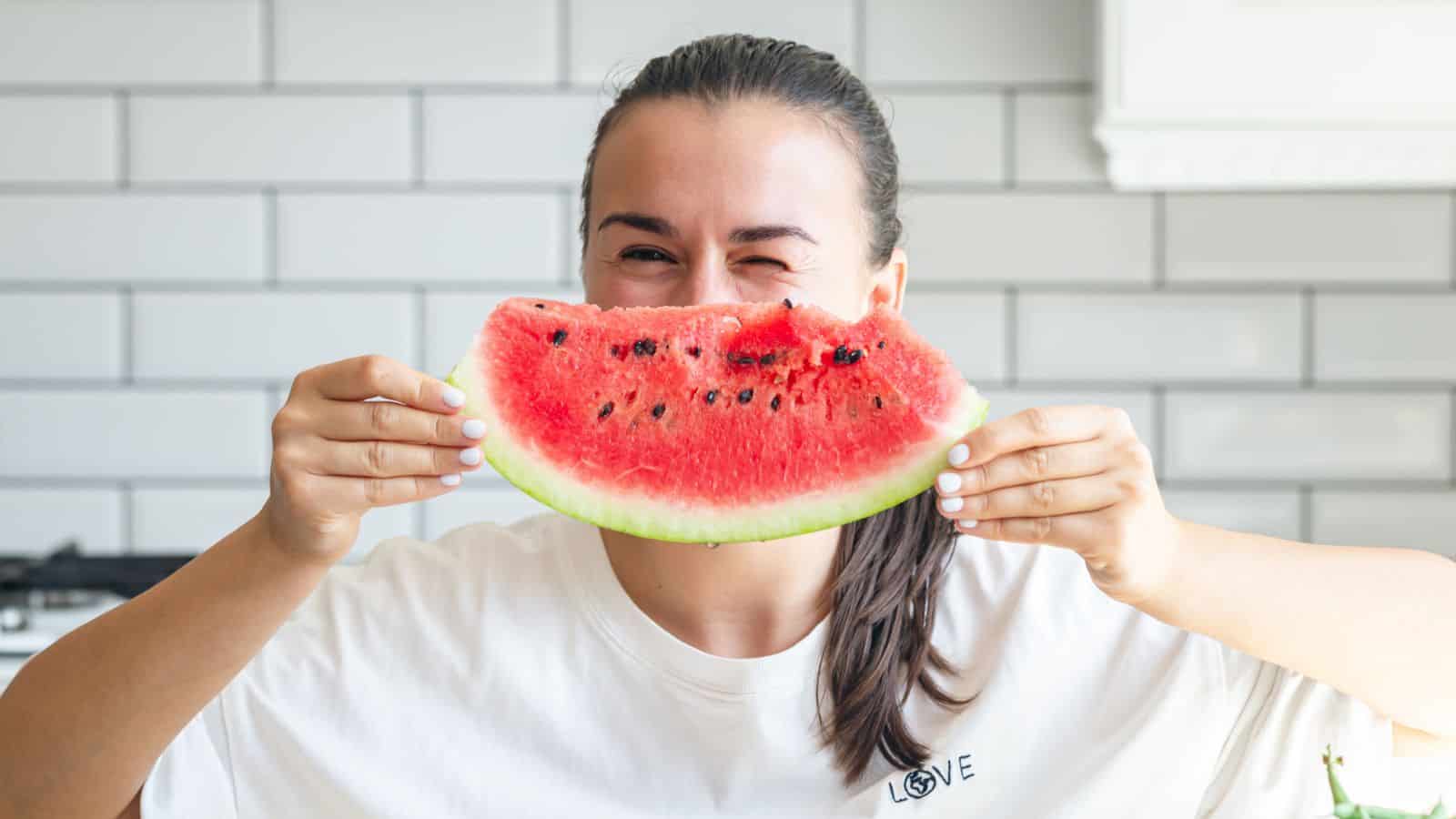 A person in a white t-shirt holds a large slice of watermelon in front of their mouth, smiling with one eye closed, standing in a kitchen with white tile walls.