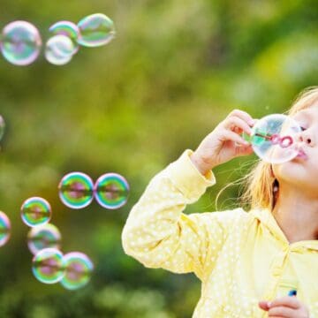 A young child in a yellow polka-dot hoodie blows soap bubbles outdoors. The background is green and blurred, with multiple bubbles floating in the air.
