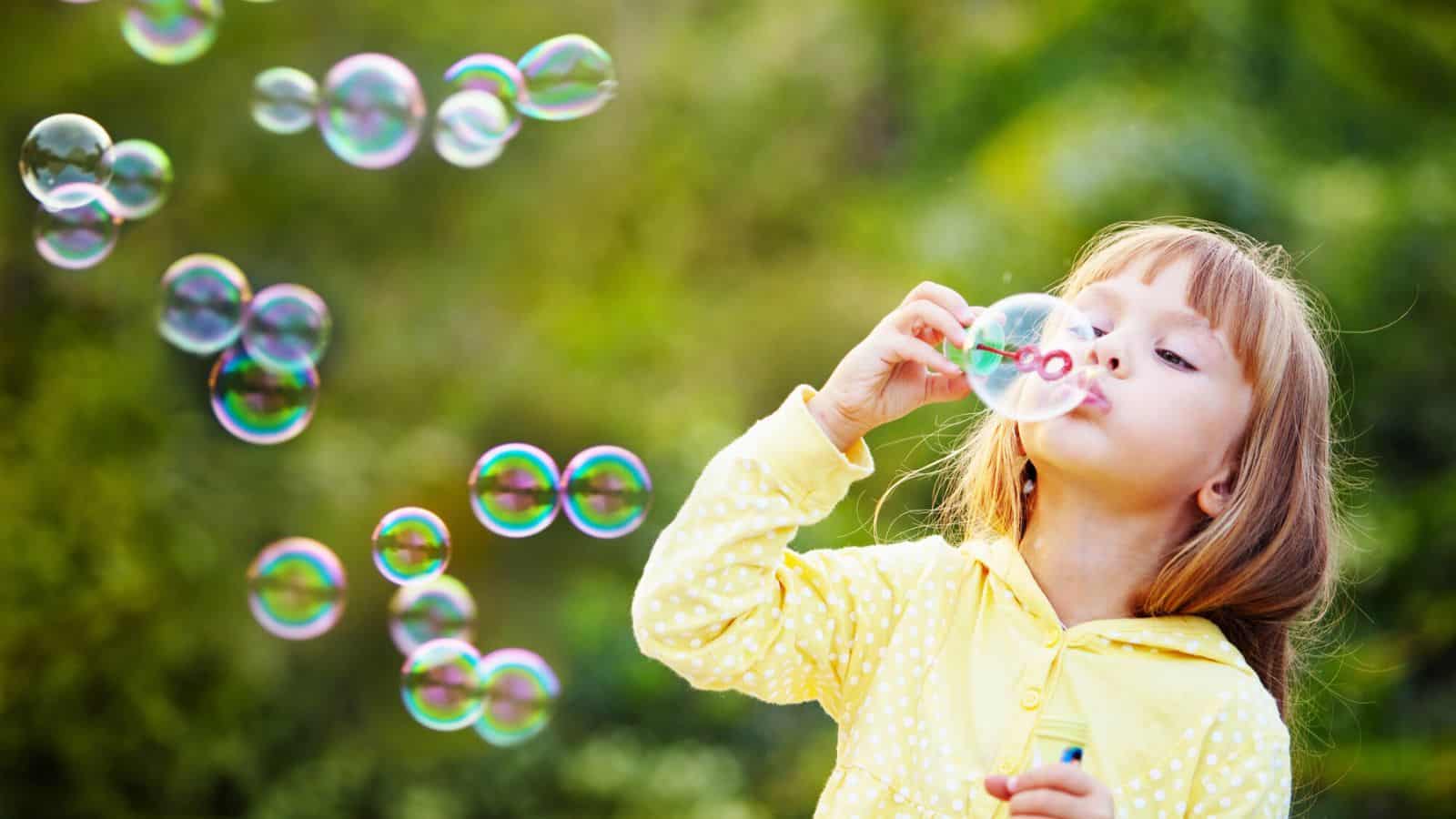 A young child in a yellow polka-dot hoodie blows soap bubbles outdoors. The background is green and blurred, with multiple bubbles floating in the air.