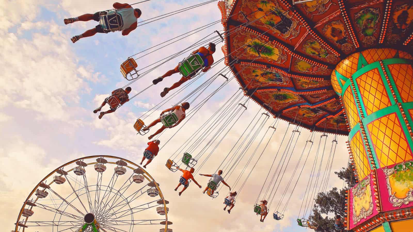 People ride a colorful swing carousel at a fairground, with legs outstretched as the ride spins. A Ferris wheel is visible in the background against a partly cloudy sky.