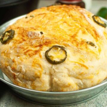 A round loaf of bread with a golden crust, topped with melted cheese and sliced jalapeños, sits in a metal baking dish on a light-colored surface. A blue-striped cloth is partially visible on the left.