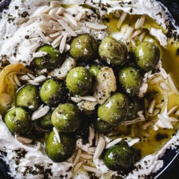 A bowl of whipped labneh topped with green olives, olive oil, slivered almonds, and herbs, surrounded by fresh parsley leaves.