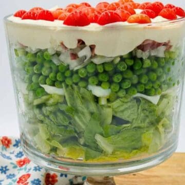 A large glass trifle bowl filled with layered salad ingredients, including lettuce, peas, chopped onions, a creamy layer, and halved cherry tomatoes on top. The bowl is on a wooden board with a floral cloth beside it.