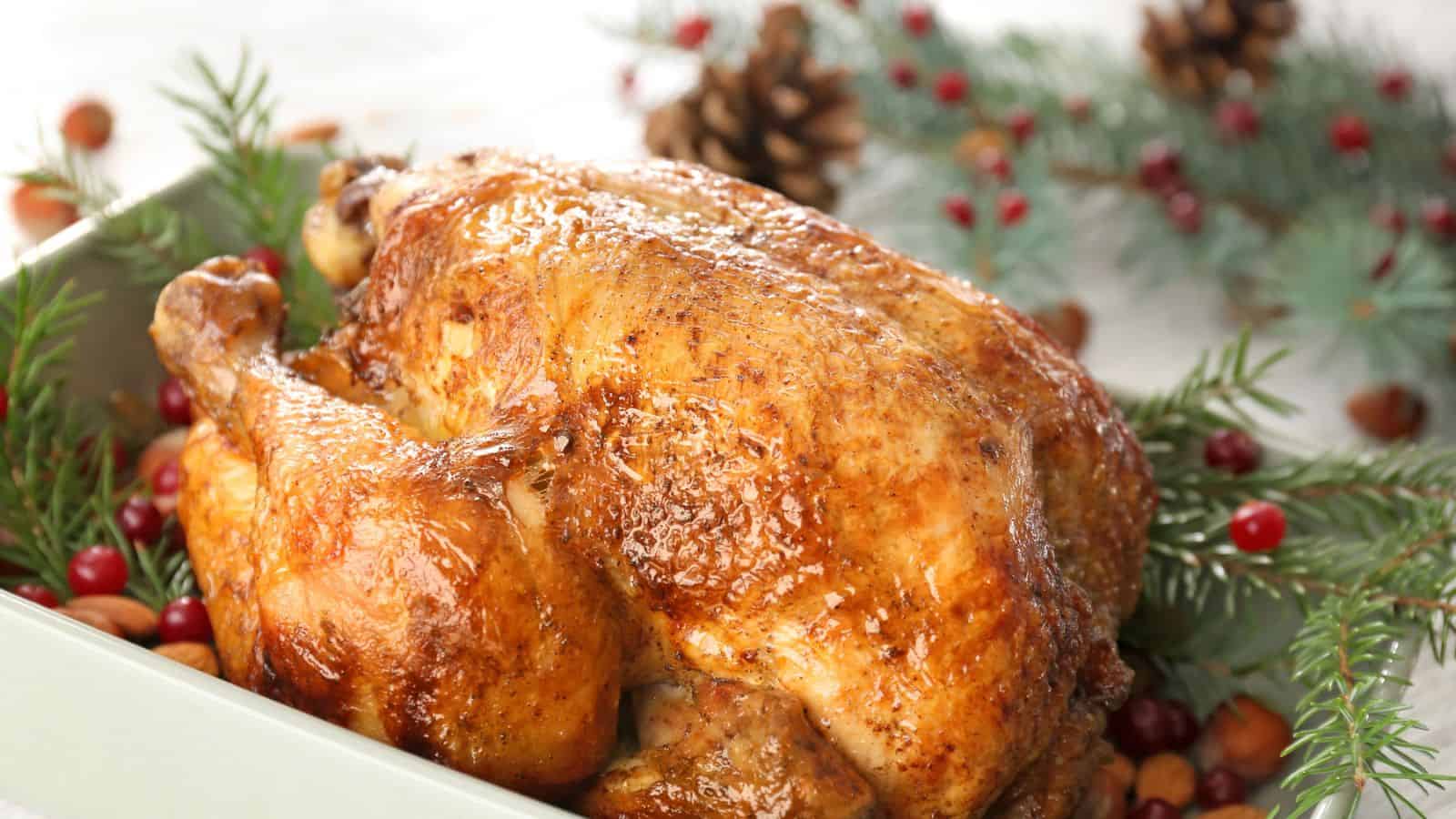 A whole roasted chicken with golden, crispy skin sits in a dish, surrounded by pine branches, pinecones, and red berries, suggesting a holiday or winter setting.