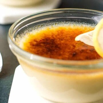 A glass ramekin filled with crème brûlée topped with a caramelized sugar layer and garnished with two thin lemon slices, placed next to a metal spoon on a dark surface.