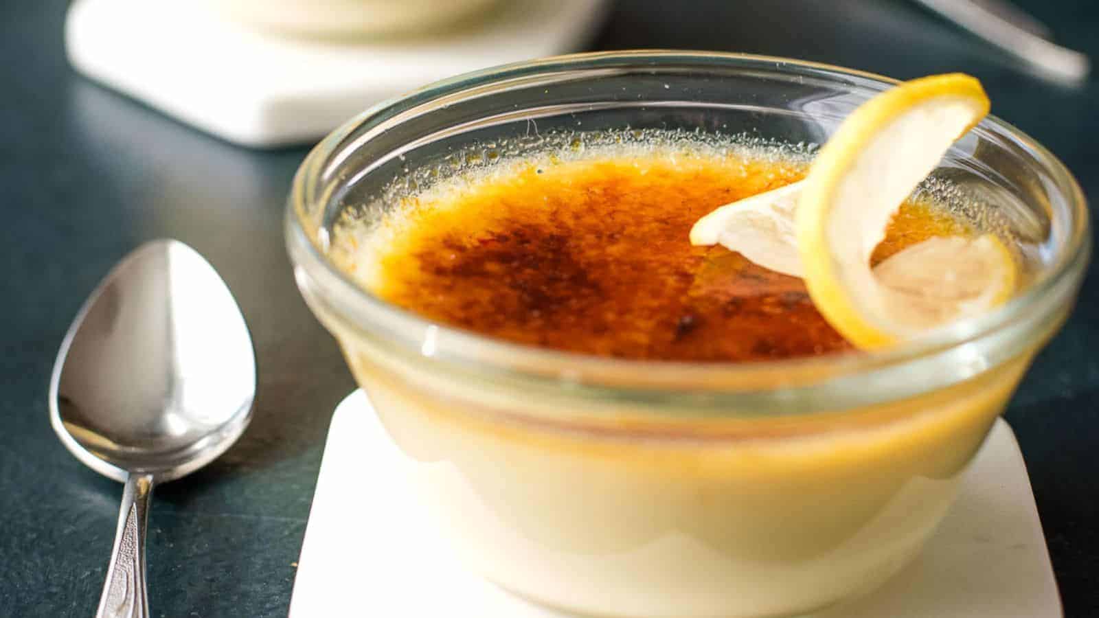 A glass ramekin filled with crème brûlée topped with a caramelized sugar layer and garnished with two thin lemon slices, placed next to a metal spoon on a dark surface.