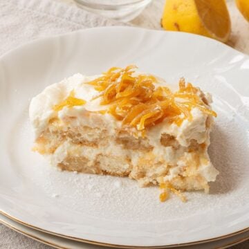 A slice of creamy dessert topped with whipped cream and thin strips of candied lemon zest sits on a white plate. The layered dessert appears soft and slightly crumbly. A lemon is partially visible in the background.