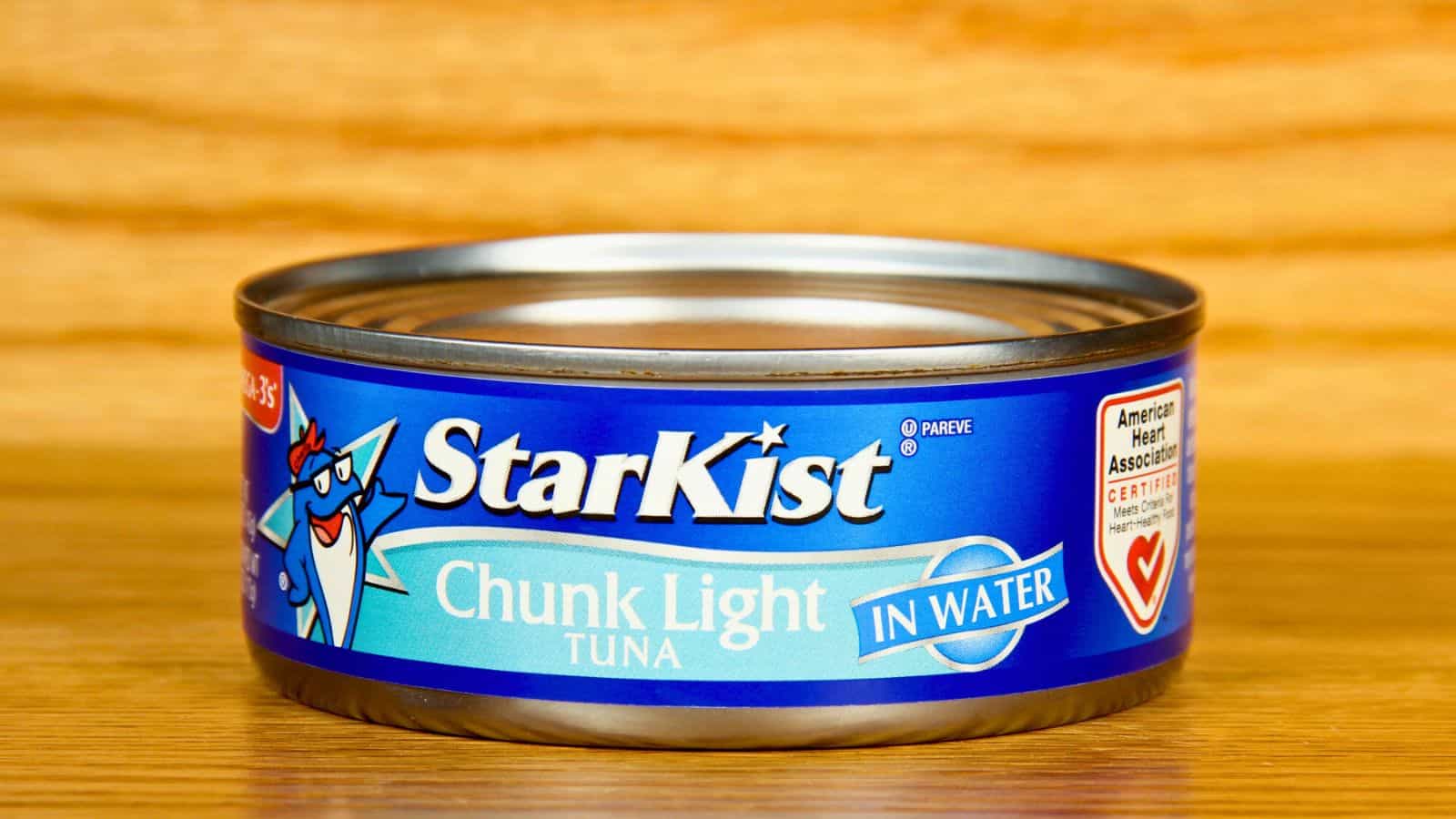 A can of StarKist Chunk Light Tuna in water sits on a wooden surface. The blue label features a cartoon fish and American Heart Association certification logo.