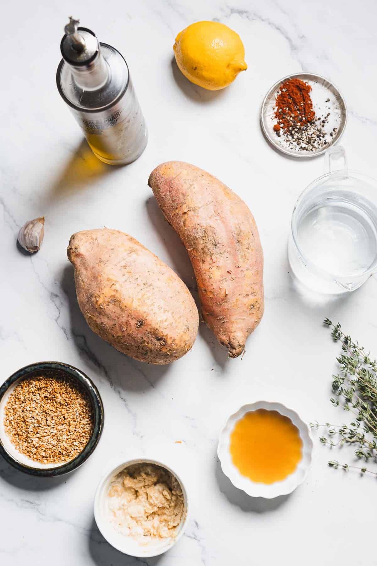 Two whole sweet potatoes on a white marble surface surrounded by a lemon, olive oil, water, garlic, fresh thyme, spices, tahini, sesame seeds, and a small bowl of maple syrup—perfect for Maple Tahini Sweet Potatoes.