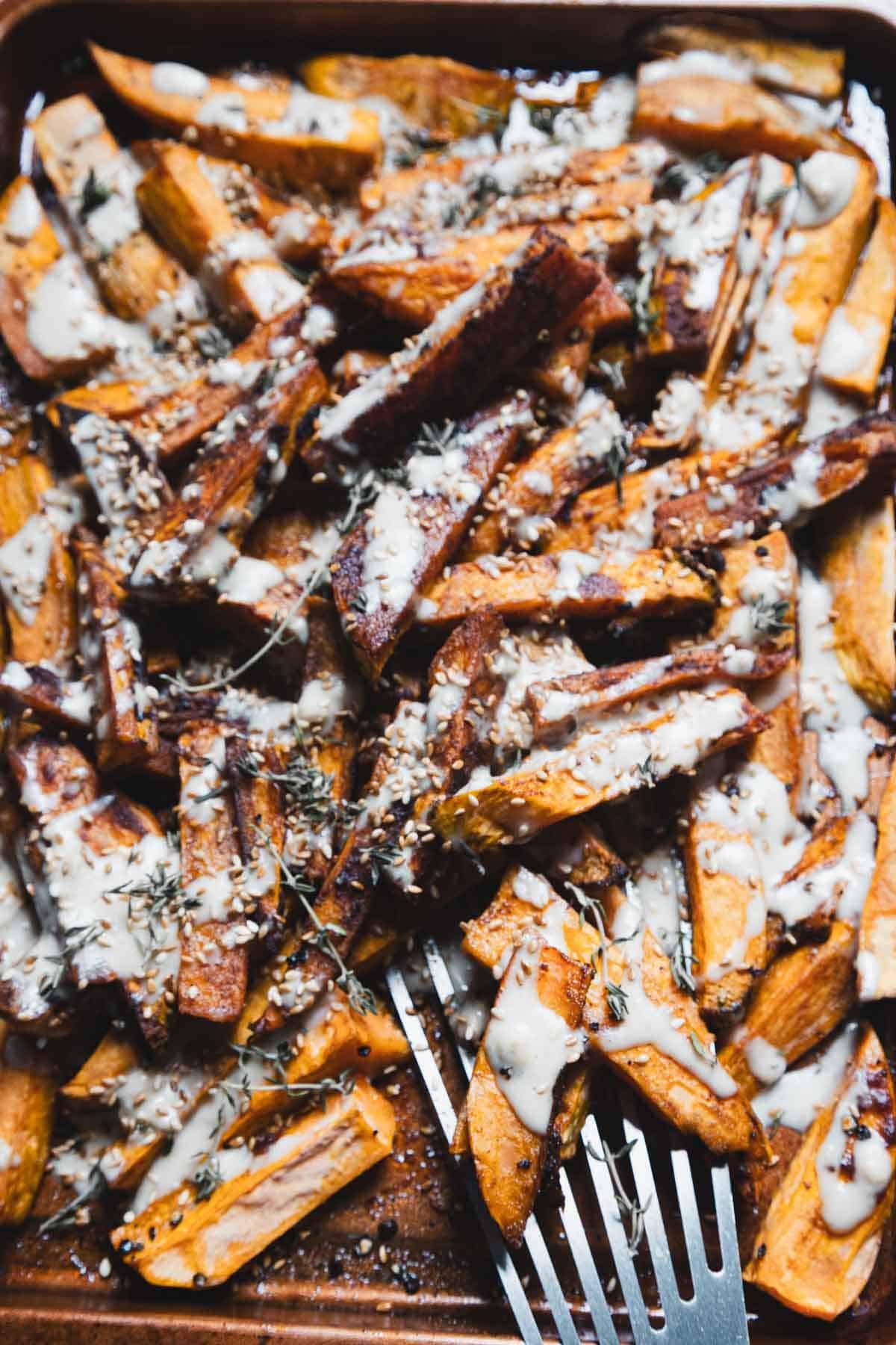 A close-up of Maple Tahini Sweet Potatoes on a baking sheet—roasted sweet potato fries drizzled with creamy white sauce, sprinkled with herbs and seeds, with a metal serving fork nestled among the fries.