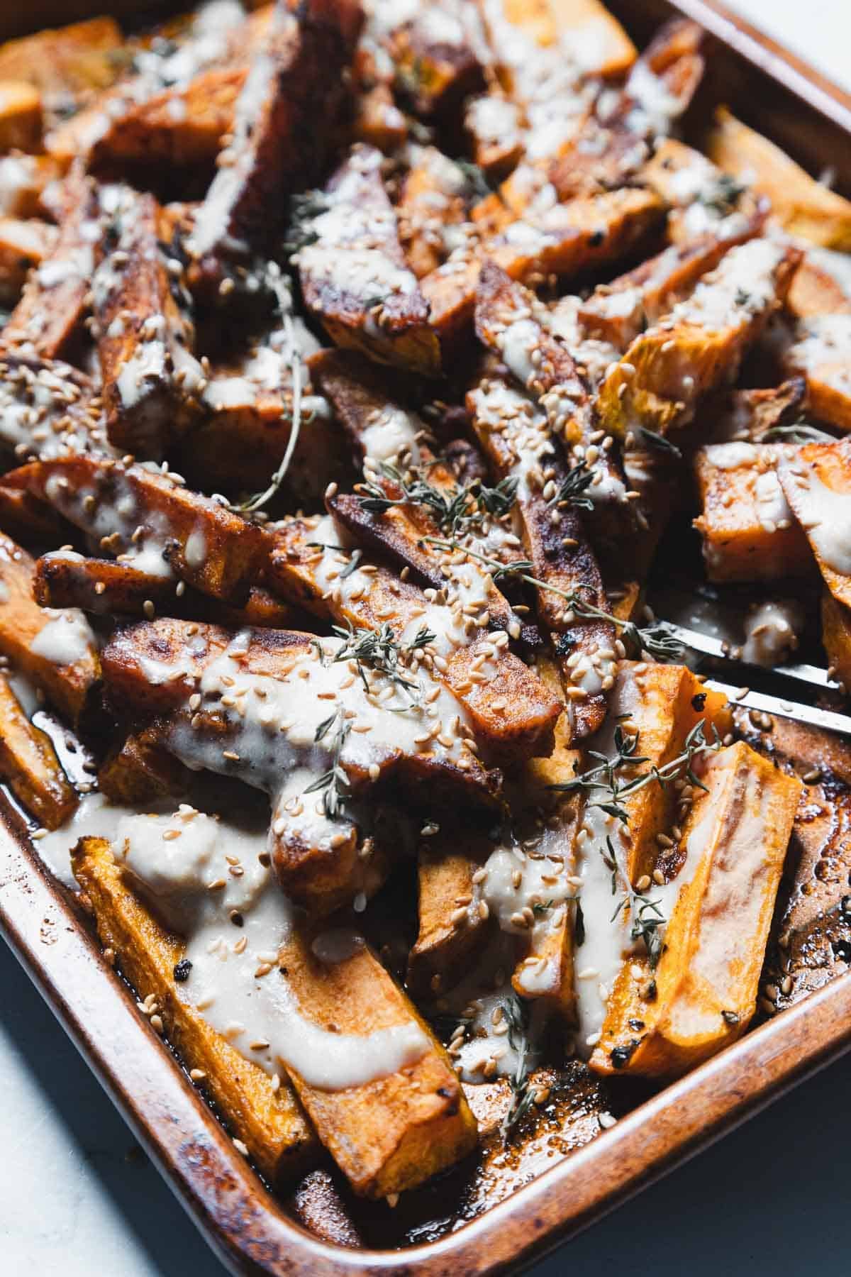 Baked sweet potato fries on a baking sheet, topped with a drizzle of creamy sauce and sprinkled with seeds and fresh herbs. A serving fork is visible on the right side of the tray.