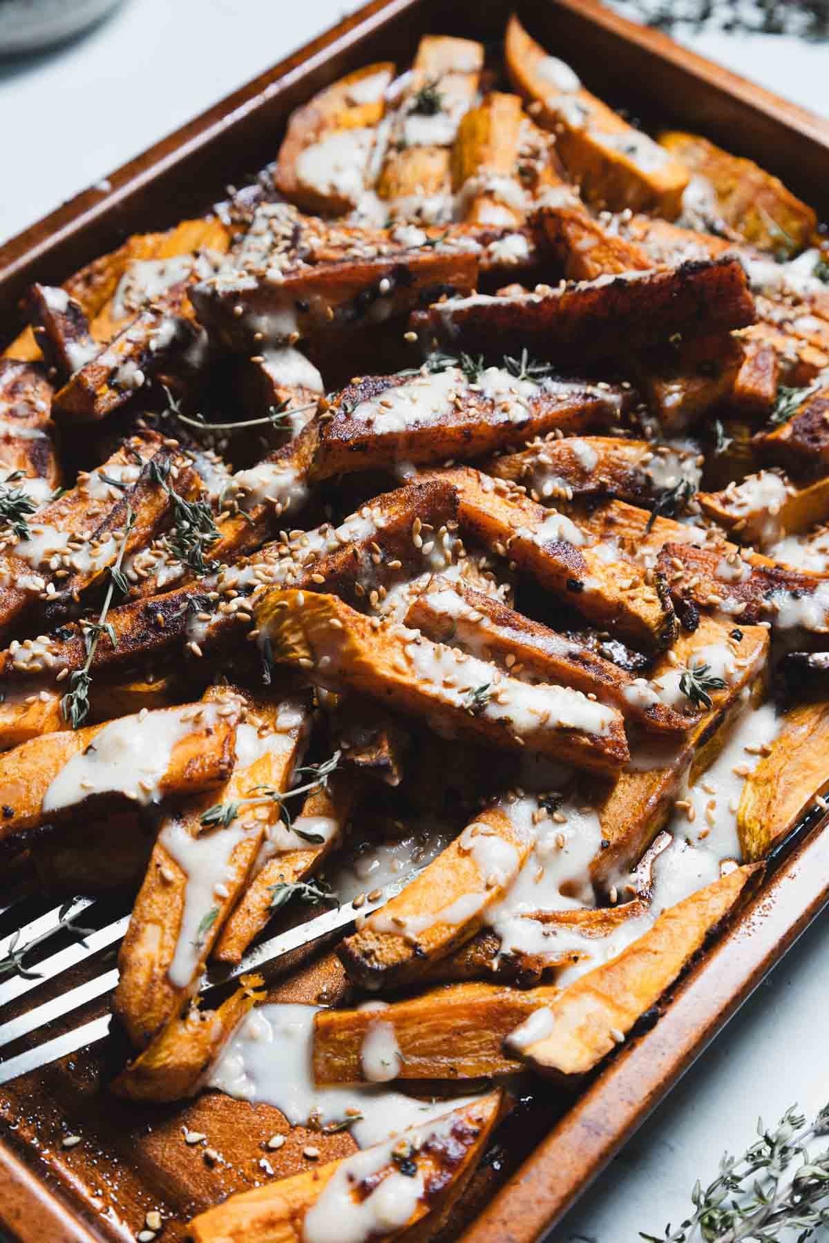 A tray of Maple Tahini Sweet Potatoes, roasted to perfection and topped with creamy white sauce, sesame seeds, and fresh herb sprigs. A metal fork rests on the tray among the wedges.