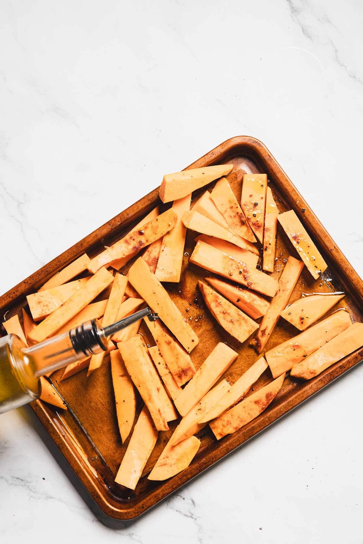A baking sheet with uncooked sweet potato fries, cut into thick strips and sprinkled with seasoning. A bottle of olive oil is being poured over these Maple Tahini Sweet Potatoes. The background is a white marble surface.