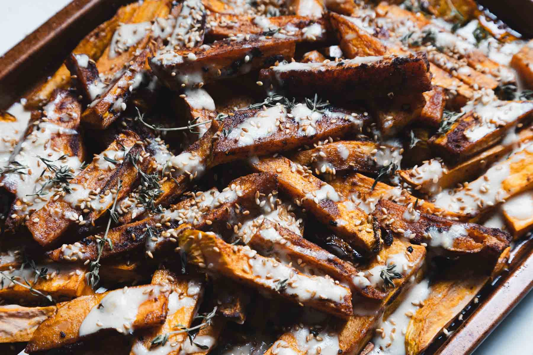 A baking sheet filled with Maple Tahini Sweet Potatoes—roasted sweet potato wedges topped with a creamy white sauce, sesame seeds, and fresh herbs.